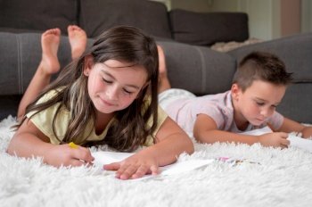 Siblings playing together at home. little boy and girl lying on the carpet and drawing on white sheets of paper with colorful crayons. High quality photography.. Siblings playing together at home. little boy and girl lying on the carpet and drawing on white sheets of paper with colorful crayons