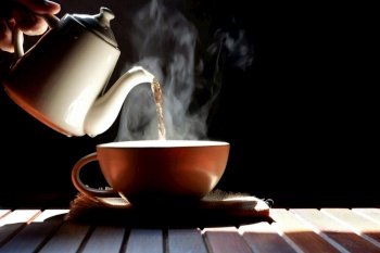 Man’s hand pouring hot tea from ceramic teapot into tea cup with steam on wooden table in dark vintage tone style