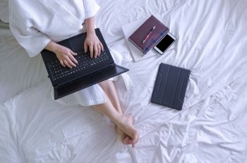 Top view of woman in white bathrobe using laptop computer with book and various device on bed in her bedroom