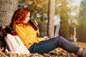 Young woman drinks coffee and works using laptop in nature. Remote work on the Internet. Rest and relaxation on an autumn sunny day.. Young woman drinks coffee and works using laptop in nature. Remote work on the Internet. Rest and relaxation on autumn sunny day.