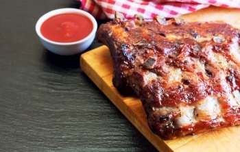 grilled pork ribs on a wooden cutting board with tomato ketchup on black slate surface. grilled pork ribs on a cutting board