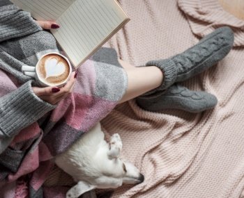 Cozy home, woman covered with warm blanket, drinks coffee,  sleeping dog next to woman. Relax, carefree, comfort lifestyle.