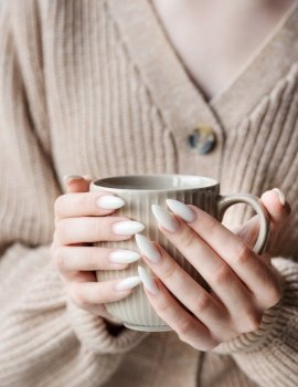 Beautiful hands of a young woman with white manicure on nails. Girl in a sweater holding a mug of tea