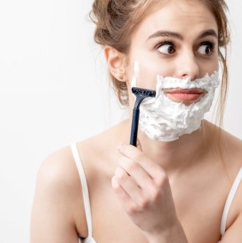 Beautiful young caucasian woman shaving her face by razor on white background. Pretty woman with shaving foam on her face. Woman shaving her face by razor