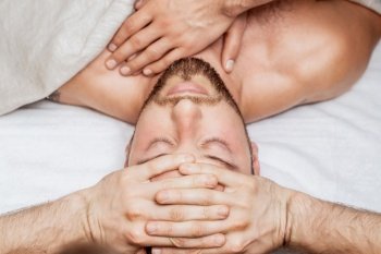 Handsome young man receiving relaxing head massage by four hand of two masseurs in health spa center. Man receiving head massage by two masseurs