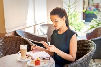 Young business woman makes notes in notebook from smartphone looking at it working at coffee break in cafe. Business woman makes notes in notebook