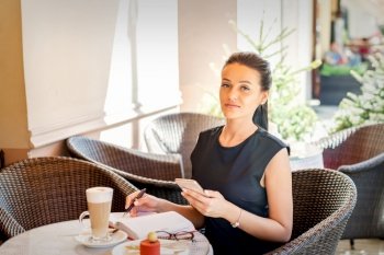 Portrait of young business woman makes notes in notebook from smartphone working at coffee break in cafe. Business woman makes notes in notebook