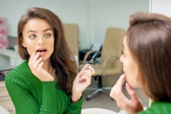 Beautiful young caucasian woman applying gloss to the lips by finger looking in the mirror. Woman applying gloss to the lips