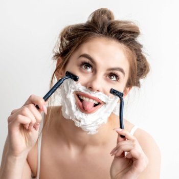Beautiful young caucasian woman shaving her face by two razors with tongue out on white background.. Woman with shaving foam on her face
