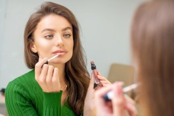 Beautiful young caucasian woman applying gloss to the lips looking in the mirror. Woman applying gloss to the lips