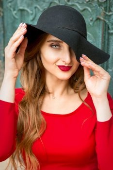 Portrait of beautiful fashionable caucasian young woman in red dress wearing black hat looking at camera. Woman in red dress wearing black hat