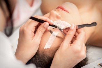 Young caucasian woman having eyelash extension procedure in beauty salon. Beautician glues eyelashes with tweezers. Woman having eyelash extension procedure