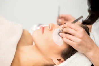 Young caucasian woman having eyelash extension procedure in beauty salon. Beautician glues eyelashes with tweezers. Woman having eyelash extension procedure
