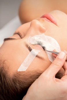Closeup view of young caucasian woman having eyelash extension procedure in beauty salon. Beautician glues eyelashes with tweezers. Woman having eyelash extension procedure