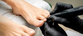 Pedicure master removes cuticle from toes of woman using professional electric nail hardware in nail salon. Pedicurist removes cuticle from toes