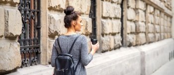 Back view of a young woman with a hot drink and backpack walks in a European city. Woman with hot drink and backpack