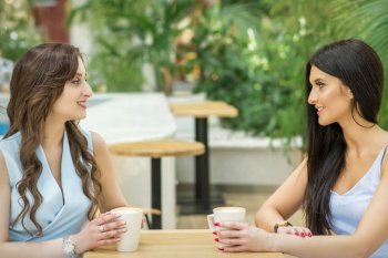 Two beautiful young caucasian women drink coffee sitting at the table in cafe outdoor. Two young women drink coffee