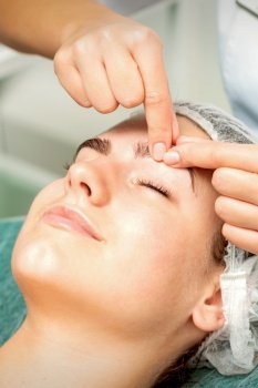 Beautician’s hands make massage on eyebrow for a woman while a facial massage in cosmetology clinic center. Beautician make massage on eyebrow