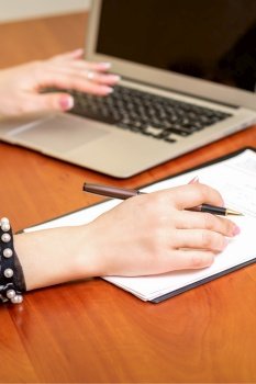 Female hands holding pen under the document and working with laptop at the table in an office. Female working with the documents