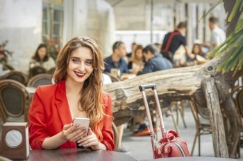 Beautiful young travel woman with smartphone sitting at the table looking at camera in cafe outdoors. Woman with smartphone sitting in cafe