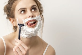 Beautiful young caucasian smiling woman shaving her face with razor sticking out tongue looking up on white background. Woman shaving face with razor