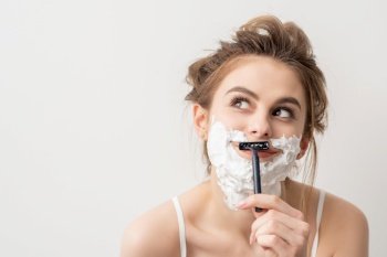 Beautiful young caucasian smiling woman shaving her face with razor looking up on white background. Woman shaving face with razor