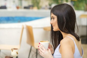 The side view of a young woman drinking coffee resting at the resort at a cafe outdoors. The side view of a young woman drinking coffee resting at the resort at a cafe outdoors.