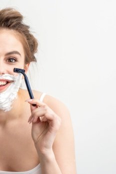 Beautiful young caucasian woman shaving her face by razor on white background. Pretty smiling woman with shaving foam and razor on her face. Beautiful young caucasian woman shaving her face by razor on white background. Pretty smiling woman with shaving foam and razor on her face.