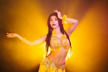 Seductive sexy traditional oriental belly dancer girl dancing on yellow neon smoke background. Woman in exotic costume with feathers sexually moves her semi-nude body.. Seductive sexy traditional oriental belly dancer girl dancing on yellow neon smoke background. Woman in exotic costume with feathers sexually moves her semi-nude body