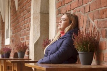 Caucasian woman in coat rests at small table in street cafe next to the brick wall in autumn