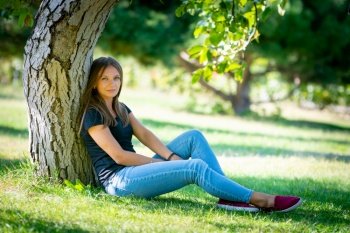 A girl sits under a tree in a sunny park and looks into the frame a. A girl sits under a tree in a sunny park and looks into the frame