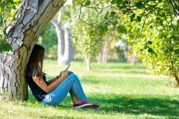 A girl sits under a tree in a sunny park and looks at the phone screen a. A girl sits under a tree in a sunny park and looks at the phone screen