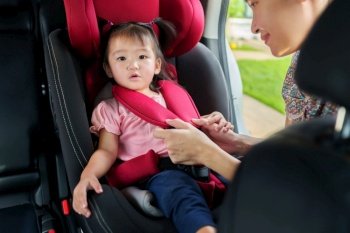 mother is fastening safety belt to toddler girl in car seat before go for ride