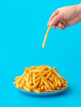 Woman hand taking french fries from a plate, minimalist on a blue background. Freshly fried potatoes on a plate in bright light on a colorful table