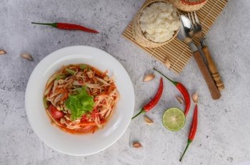 Thai papaya salad in a white plate with Sticky rice, chili, Spoon, and fork.