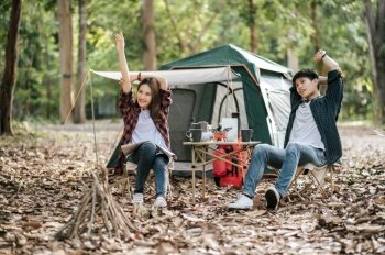 Young Asian teenager couple have relax time with camping trip, They are sitting on chair and raise up hand over head for stretching muscle at front of camping tent on backpacker trip