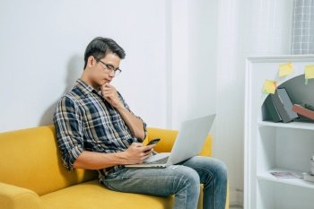 Young handsome man wearing eyeglasses with new normal, sitting on sofa and works from home on laptop computer while self-isolation and quarantine covid-19 coronavirus outbreak