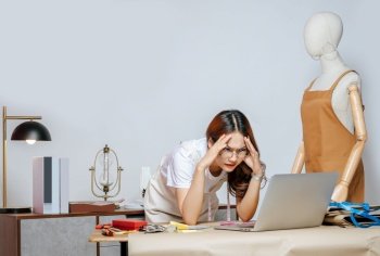 Young female fashion designer or tailor wearing eyeglasses with working with laptop computer standing with feels displeasure gesture, full of tailoring tools with mannequin and equipment on desk in the studio