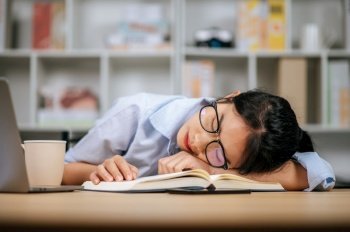 Young Asian tired woman in eyeglasses sleeping on desk with laptop and textbook at home