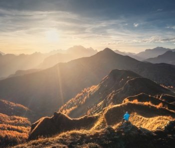 Aerial view of man on the hill and beautiful mountains in haze at colorful sunset in autumn. Dolomites, Italy. Top view of guy, mountain ridges in fog, orange grass and trees, sky in fall. Hiking. Aerial view of man on the hill and beautiful mountains in haze