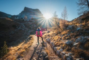 Walking girl with backpack on the trail in mountains at sunset in autumn. Beautiful landscape with young woman, high rocks, path, orange trees, sky in fall in Dolomites, Italy. Adventure and hiking. Walking girl with backpack on the trail in mountains at sunset