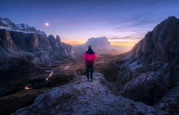 Woman on the rock and mountain peaks at night in autumn in Dolomites, Italy. Girl on the stone, high rocks, purple sky with moon, light trails on road at sunset in fall. Colorful landscape with cliffs. Woman on the rock and mountain peaks at night in autumn
