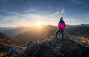 Young woman on mountain peak and beautiful mountains in haze at colorful sunset in autumn. Dolomites, Italy. Sporty girl, mountain ridges in fog, orange grass, trees, golden sun in fall. Hiking. Young woman on mountain peak and beautiful mountains