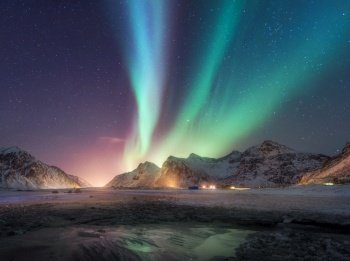Aurora borealis above the snowy mountain and sandy beach in winter at night. Northern lights in Lofoten islands, Norway. Sky with stars and polar lights. Landscape with aurora, sea coast, city lights