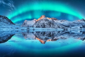 Northern lights over the snowy mountains, sea coast, reflection in water at night in Lofoten, Norway. Aurora borealis and snow covered rocks. Winter landscape with polar lights and fjord. Starry sky. Northern lights over the mountain, sea coast, reflection in water