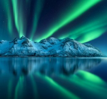 Northern lights over the snowy mountains, sea coast, reflection in water at night in Lofoten, Norway. Aurora borealis and snow covered rocks. Winter landscape with polar lights and fjord. Starry sky. Northern lights over the mountain, sea coast, reflection in water