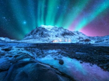 Northern lights and snowy mountains, rocky sea coast, reflection in water at night in Lofoten, Norway. Aurora borealis. Winter landscape with polar lights, city lights, houses, starry sky, beach.. Northern lights over the mountain, sea coast, city lights