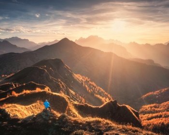 Aerial view of man on the hill and beautiful mountains in haze at sunset in autumn. Dolomites, Italy. Drone view of guy, mountain ridges in fog, orange grass, trees, sky, golden sunlight in fall