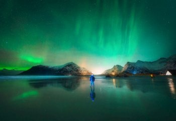 Northern Lights and man on the beach at night. Lofoten islands, Norway. Beautiful Aurora borealis. Polar lights. Winter landscape with aurora, guy, sea, starry sky, reflection in water, mountains