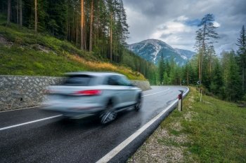Blurred car in motion on the road in green forest in rain. Dolomites, Italy. Perfect mountain road in overcast rainy summer day. Roadway, pine trees in italian alps. Transportation. Highway in wood